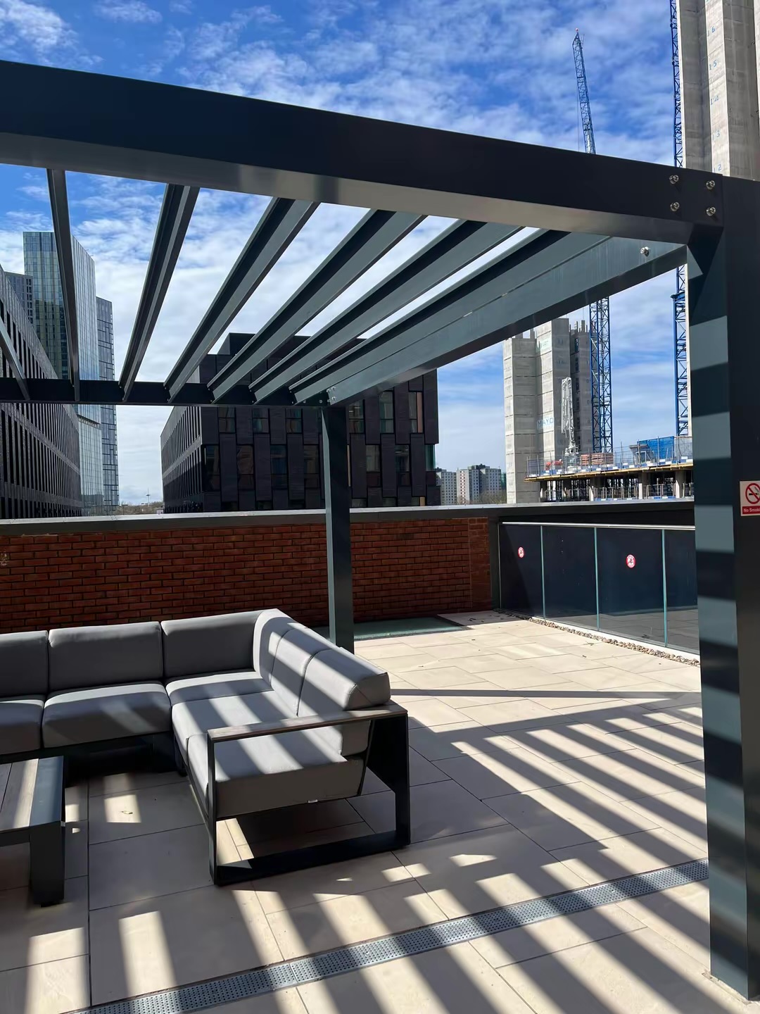 Outdoor terrace with sofa seating beneath a pergola; neighbouring construction visible beyond