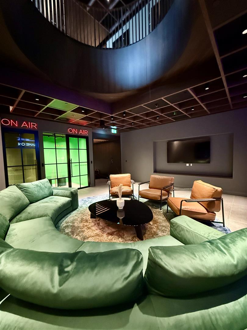 Lounge with a curved sofa and armchairs facing a wall-mounted screen, beside two glass-fronted booths with illuminated On Air signs