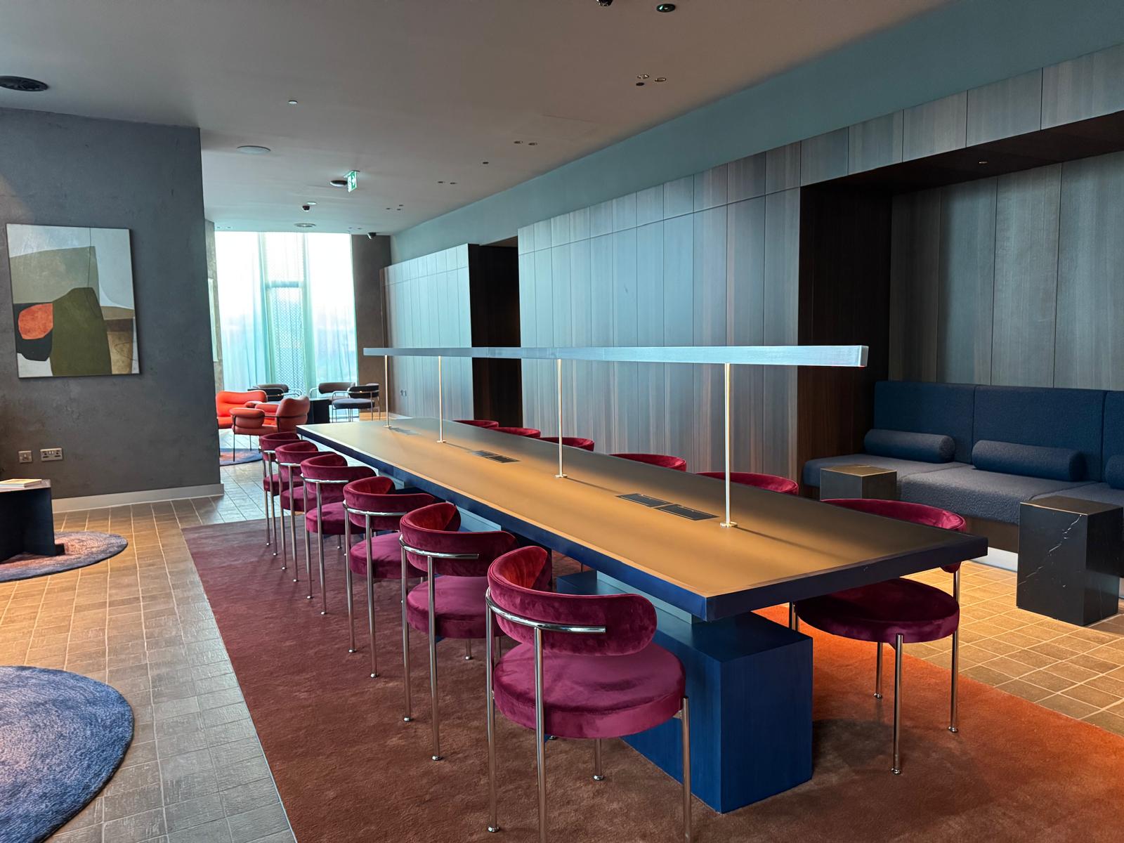 Co-working space with a long worktable, overhead linear lighting, magenta chairs and a built-in blue bench alcove