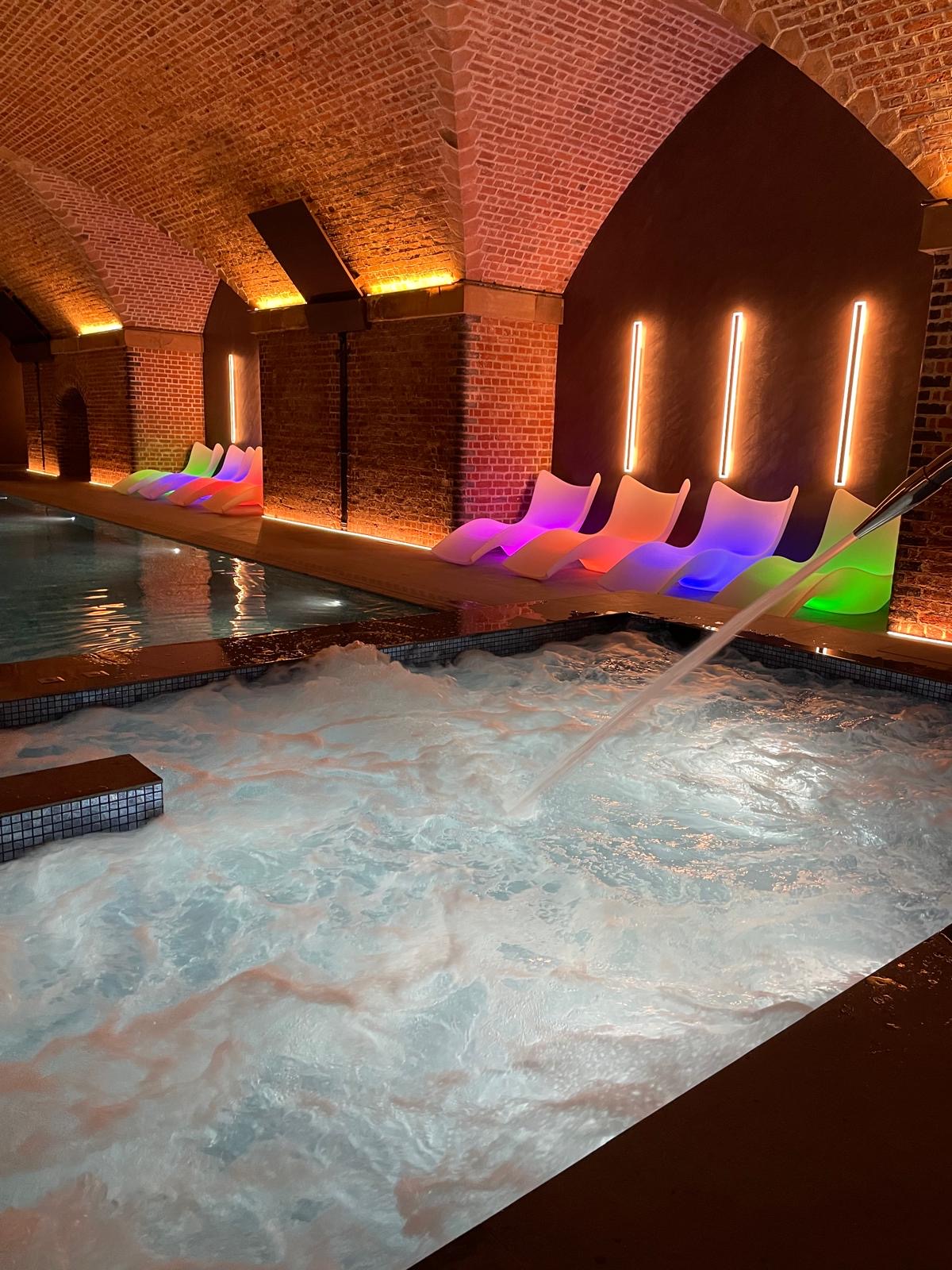 Bubbling hydrotherapy pool with a steel water spout beneath the brick arches, with illuminated loungers beside the swimming pool