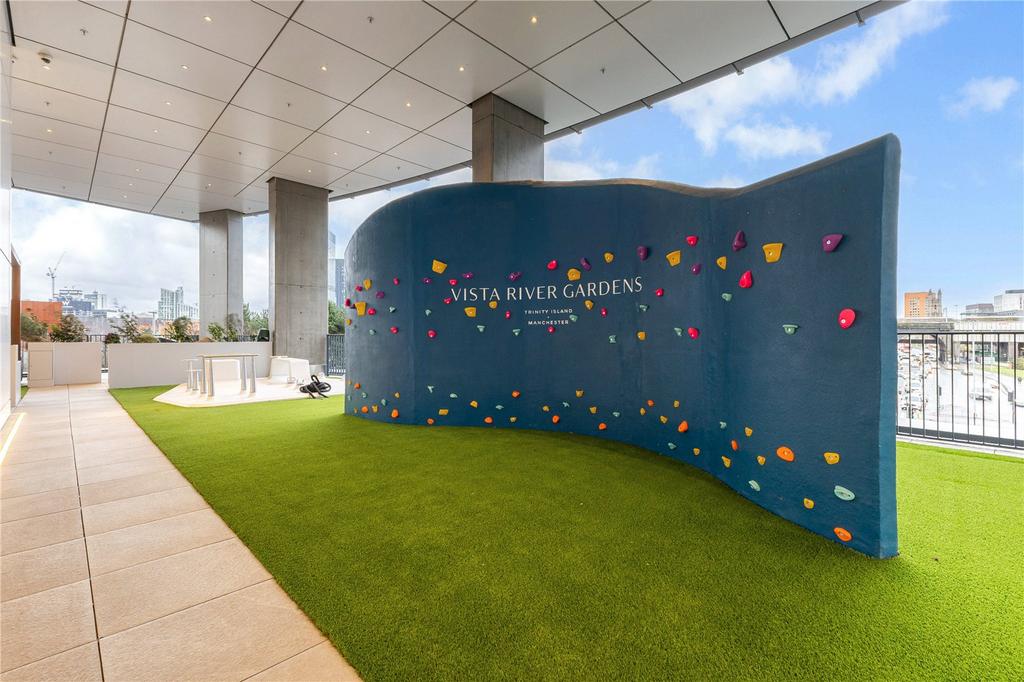 Covered outdoor terrace with a curved bouldering wall printed ‘Vista River Gardens, Trinity Island, Manchester’, artificial turf and outdoor gym equipment beyond