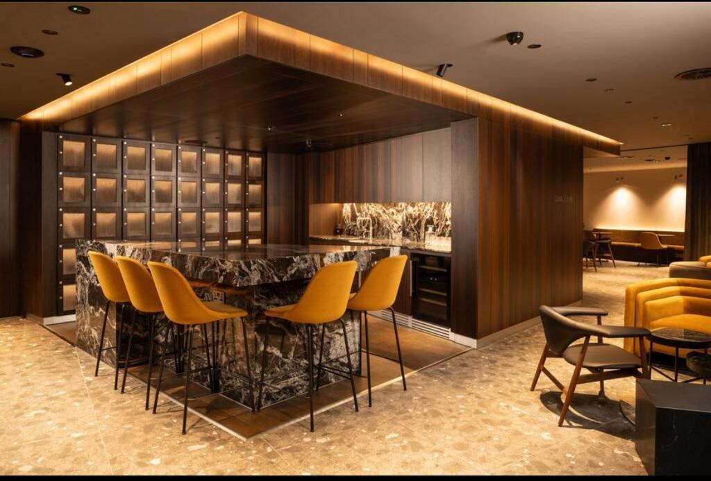 Bar area with a marble waterfall island, mustard bar stools, illuminated glass-fronted lockers and lounge seating beyond
