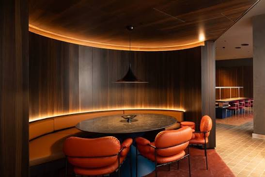Timber-panelled alcove with burnt-orange banquette seating and a round marble table, with a long communal table beyond