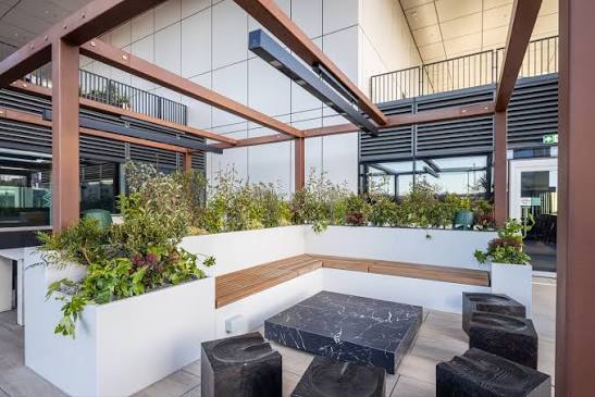 Open-air terrace with an L-shaped bench, planters, a black marble coffee table and carved block stools beneath a bronze pergola
